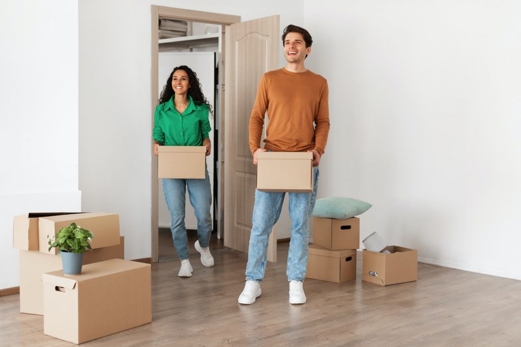 Happy couple holding cardboard boxes, walking in new house